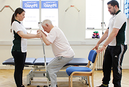 Multiple Liverpool OT staff aid male patient in standing up from chair.